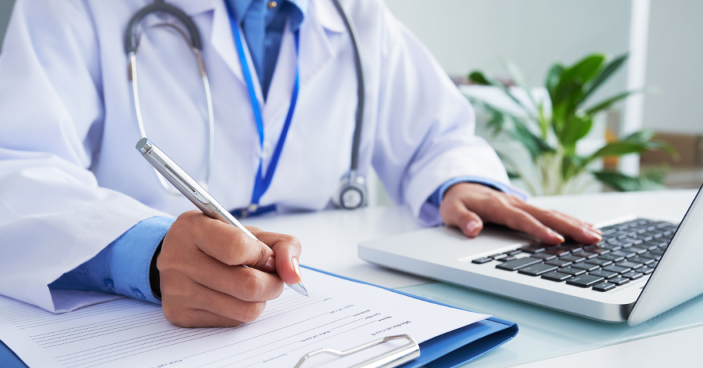 Physician sitting at desk on laptop writing information in document.